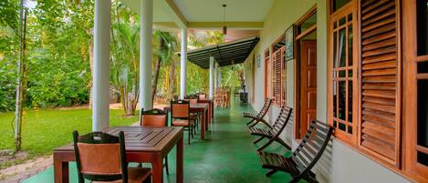A covered outdoor area with tables and chairs, surrounded by greenery and a building with wooden shutters.
