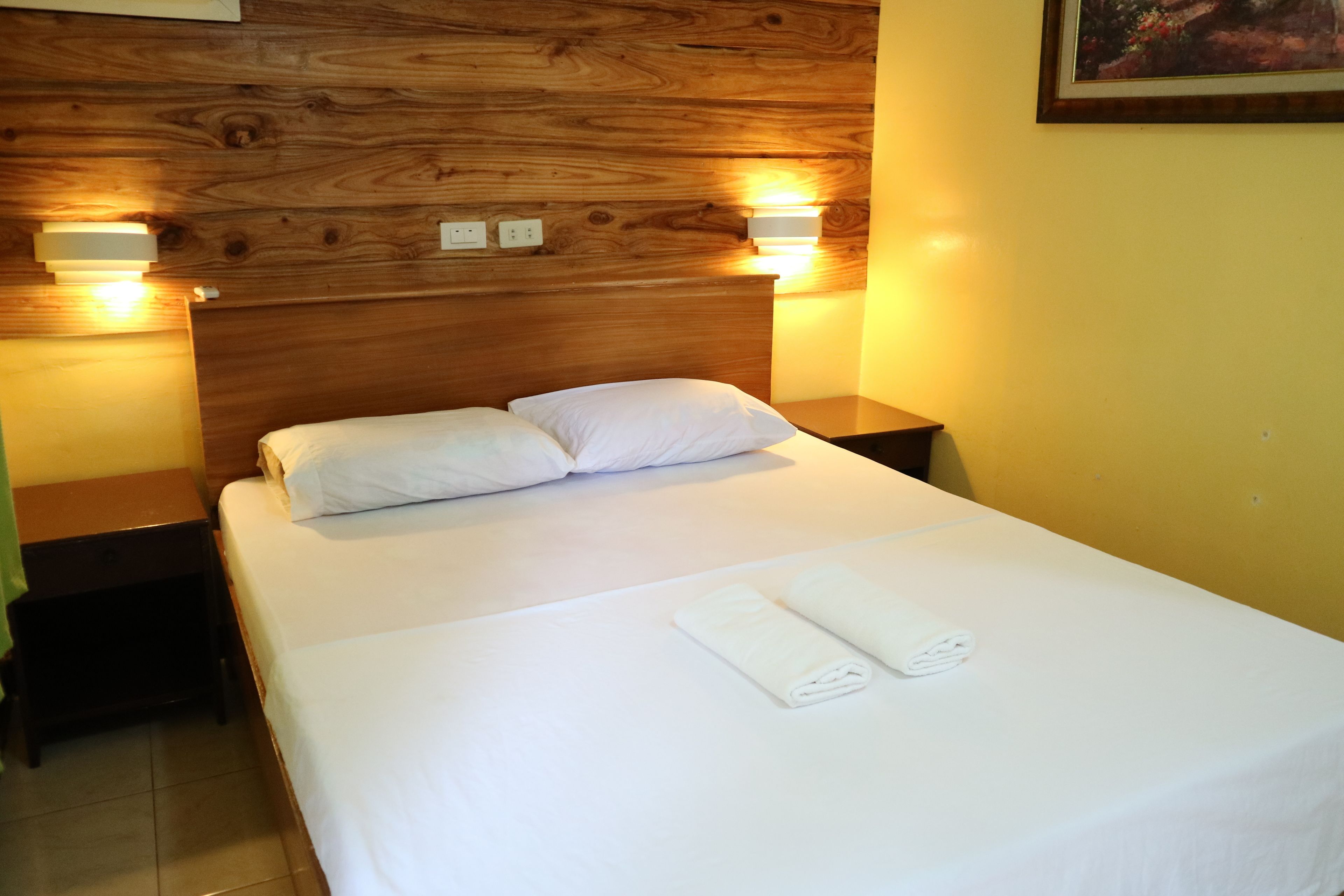 A hotel room with a wooden headboard, two bedside lamps, a bed with white linens, and a painting on the wall.