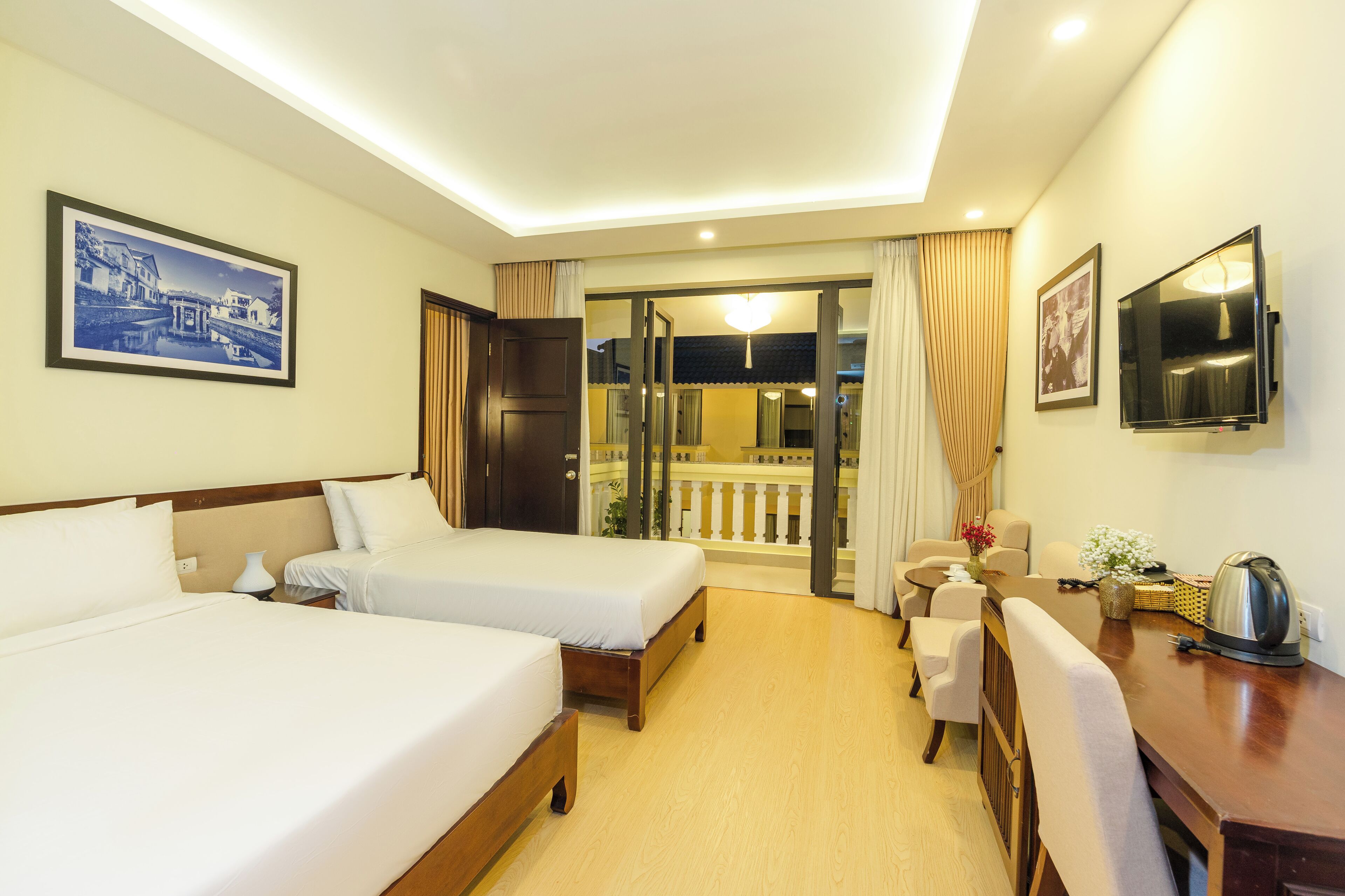 A hotel room with two beds, a TV, a desk, and a balcony with curtains.