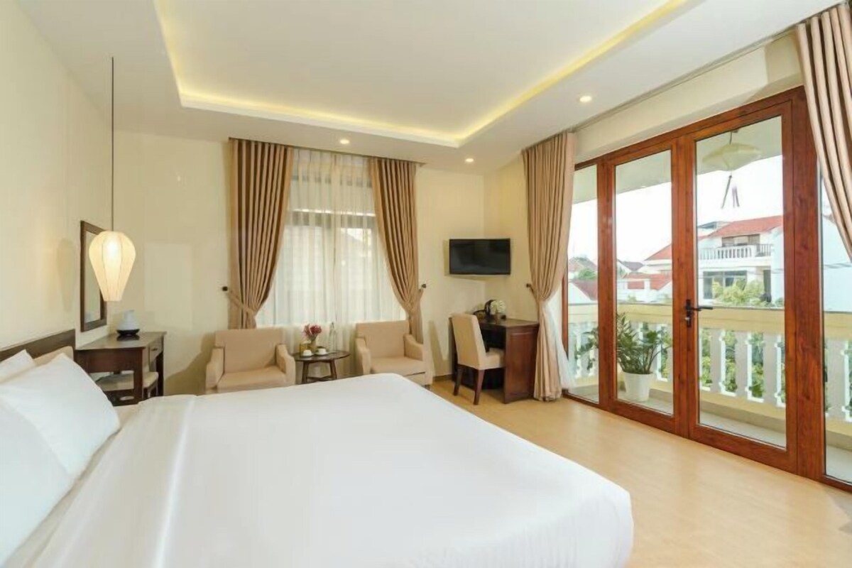 A hotel room with a large bed, a desk with a chair, a TV, and a balcony with curtains.