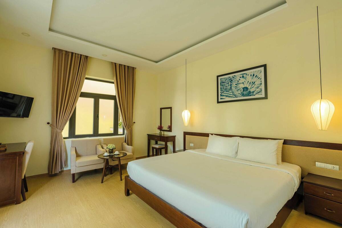 A hotel room with a large bed, a desk, a chair, a small table with a vase, and a window with curtains.
