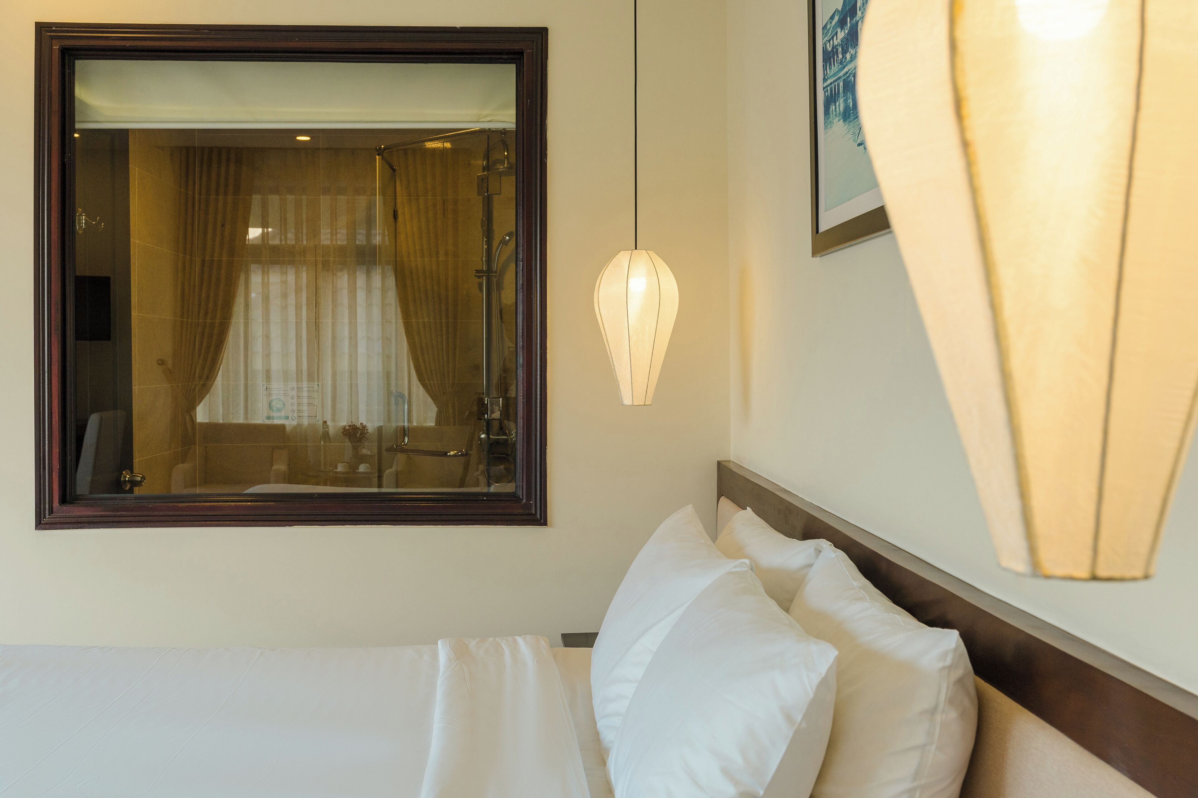 Une chambre d’hôtel avec un grand miroir, un lit recouvert de draps blancs, une lampe fixée au mur et un tableau encadré.