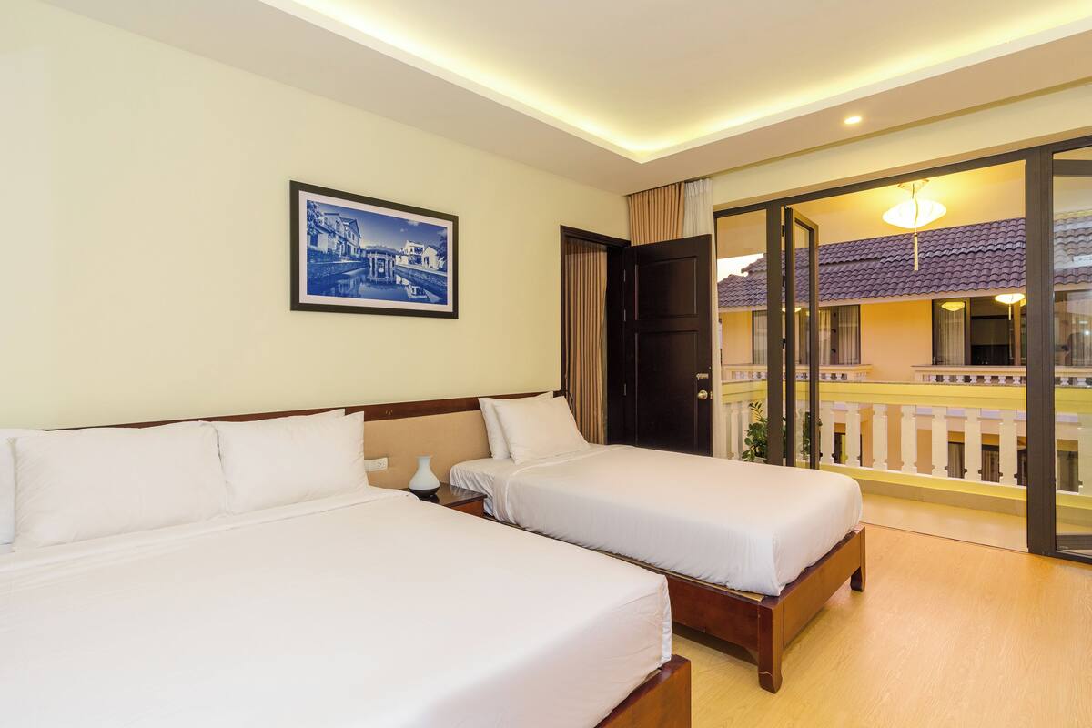A hotel room with two beds, a nightstand, a framed picture on the wall, and a balcony with a view of a building.