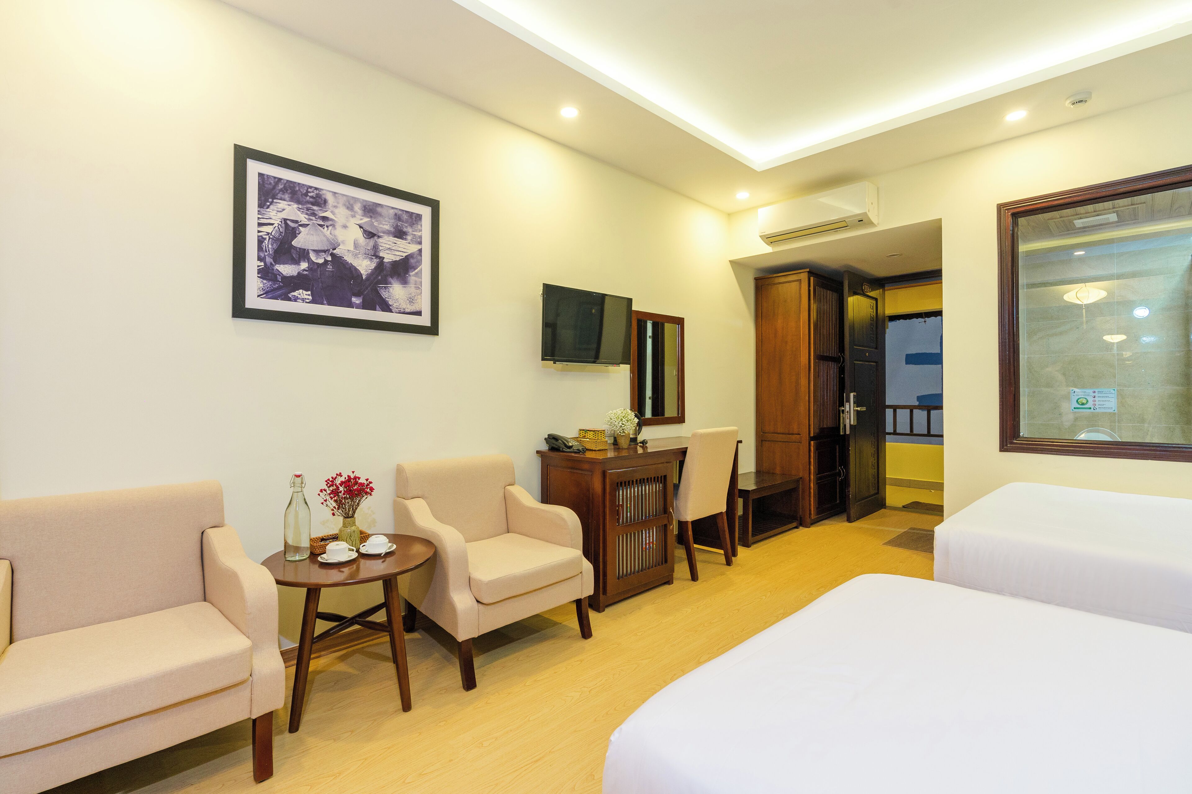A hotel room with a bed, two armchairs, a small table with a vase, a television, and a mirror.