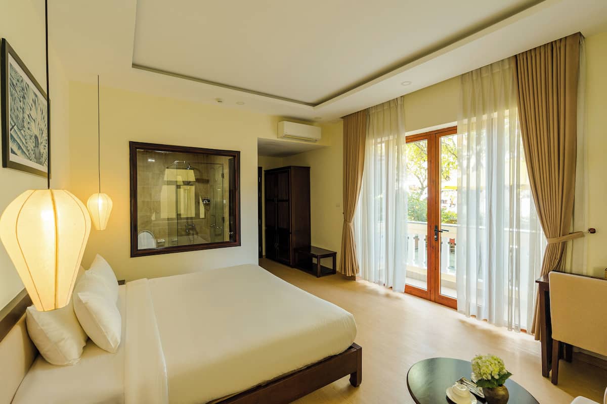 A hotel room with a large bed, a desk, a chair, and a balcony with curtains.