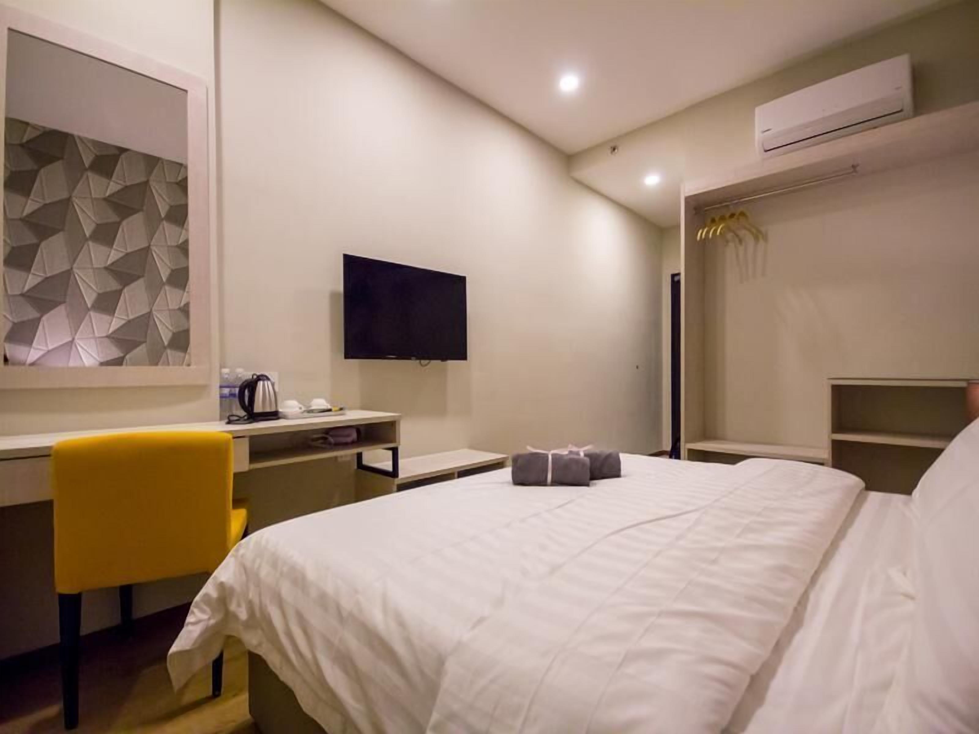 A hotel room with a bed, a desk with a chair, a flat-screen TV, a wardrobe, and a geometric-patterned wall.
