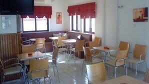 Un comedor con varias mesas y sillas, un televisor montado en la pared y ventanas amplias con cortinas rojas.