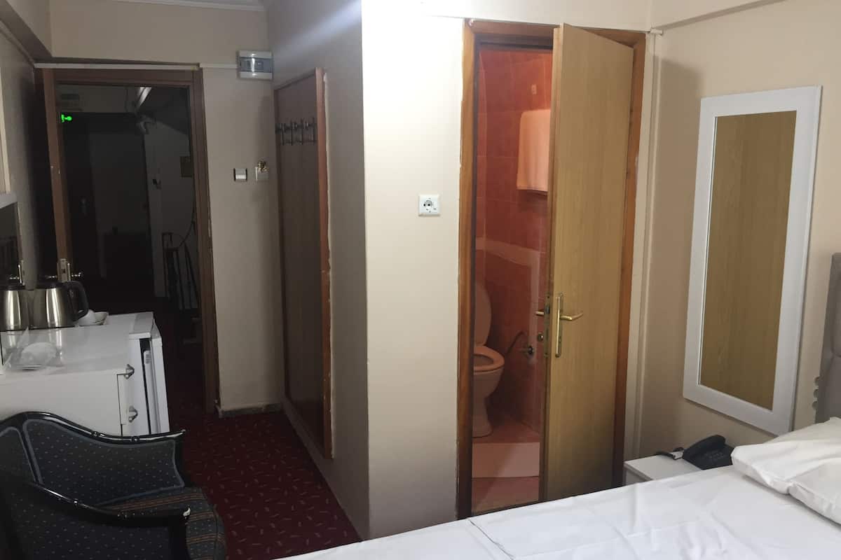 A hotel room with a bed, a desk with a kettle, a chair, a wardrobe, and a bathroom visible through an open door.