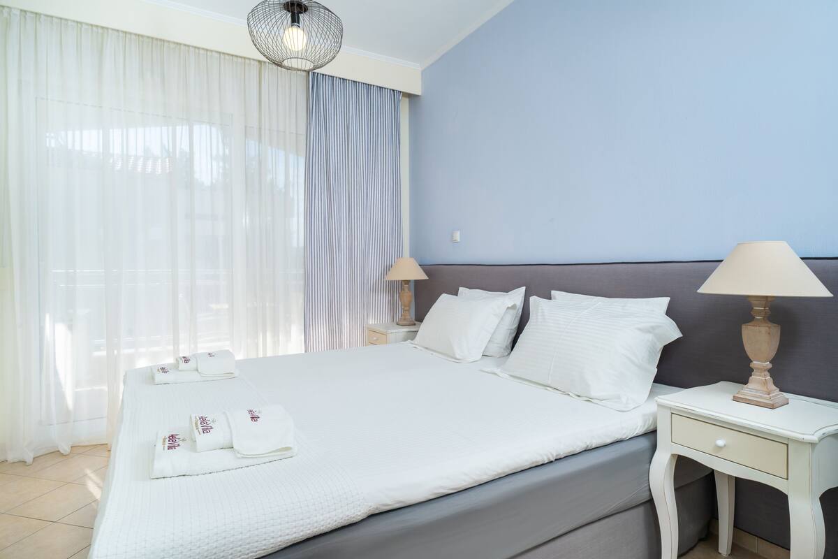 A neatly made bed with white linens and pillows, a bedside table with a lamp, and a window with sheer curtains.
