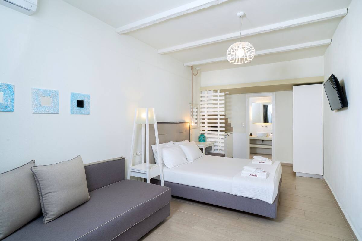 A modern hotel room with a bed, a sofa, a TV, and a bathroom visible through an open door.