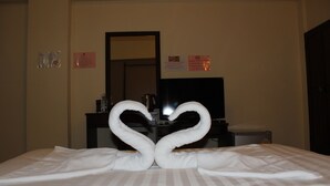 A hotel room with a bed featuring two large, heart-shaped towels. There is a television on a stand in the background, and a mirror on the wall.
