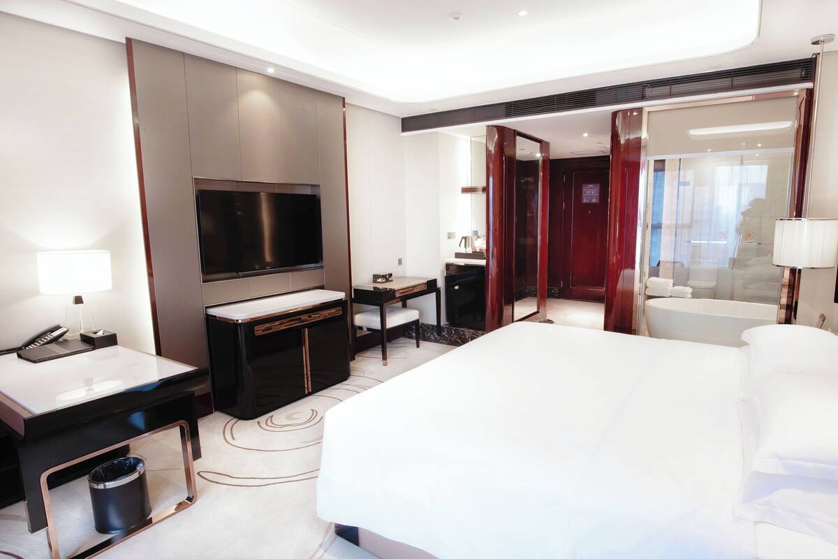Een moderne hotelkamer met een groot bed, een bureau met een flatscreen-televisie, een kaptafel met spiegel, en een aparte badkamer met bad.