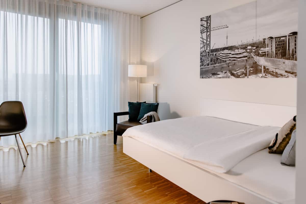A modern hotel room with a large bed, a chair, a desk, and a lamp.