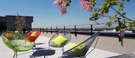 Mobiliario exterior con sillas y una mesa de madera, con vistas a un cuerpo de agua y edificios.