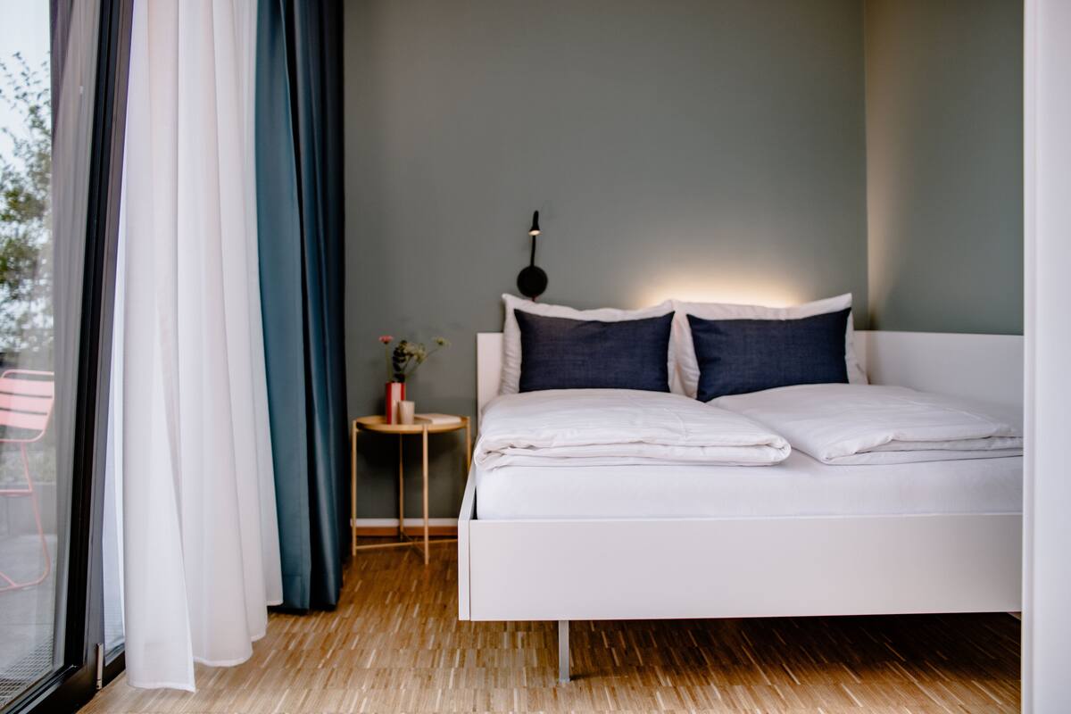 A modern bedroom with a white bed, a small bedside table with a lamp, and a view of the outdoors through a window with sheer curtains.