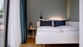 Une chambre moderne avec un lit blanc, une petite table de chevet avec une lampe, et une vue sur l’extérieur à travers une fenêtre ornée de rideaux légers.