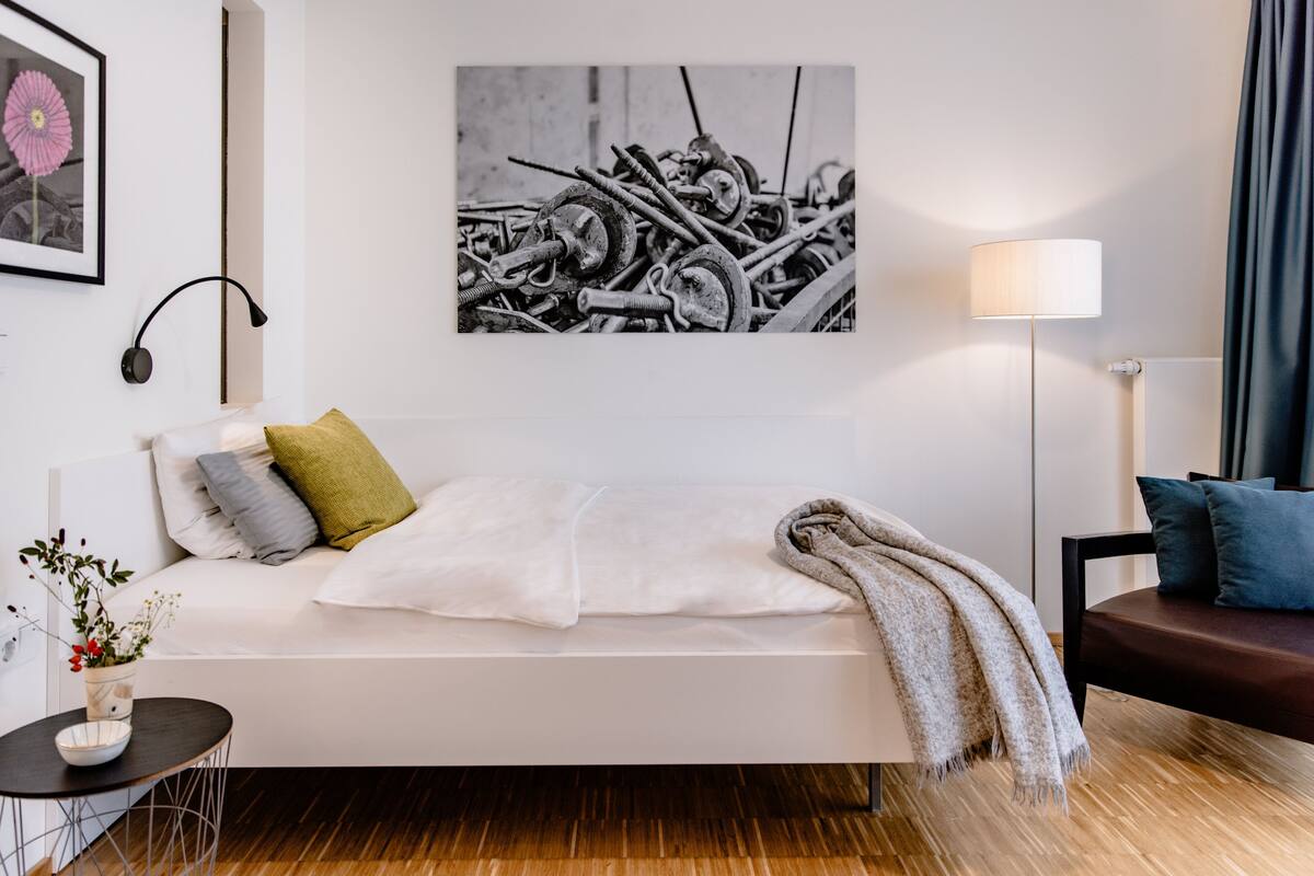 A modern bedroom with a white sofa, a small round table with a bowl of flowers, a lamp, and a framed picture on the wall.