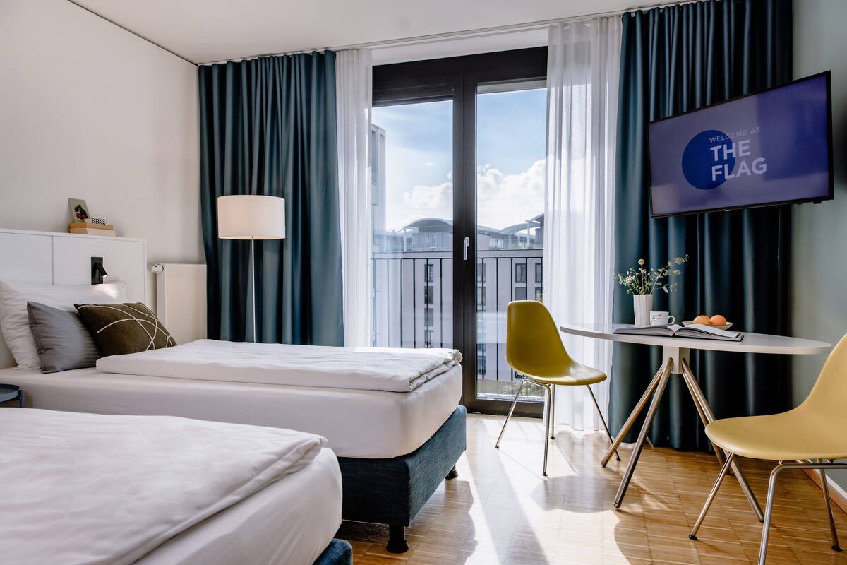 A hotel room with a bed, a desk with a chair, a TV, and a view of buildings through a large window.