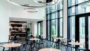 A modern dining area with round tables and black chairs, large windows, and a visible kitchen area in the background. - THE FLAG München M. (Munich)