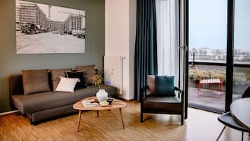 Ein modernes Wohnzimmer mit einer Couch, einem Couchtisch und einem Sessel. Es gibt ein großes Fenster mit Vorhängen, einen Blick auf die Stadt und ein gerahmtes Bild an der Wand.