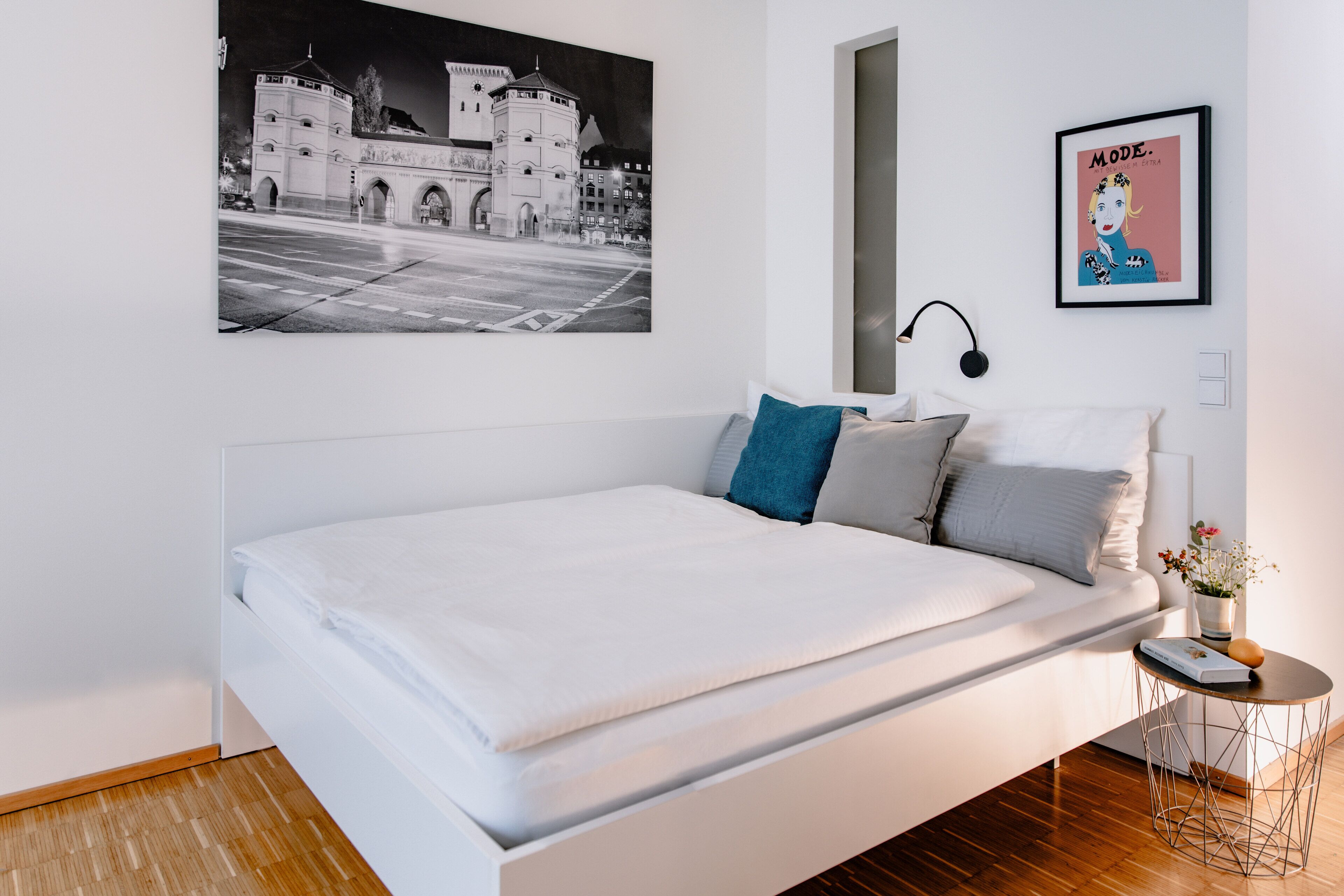A modern bedroom with a white bed, a nightstand, a framed picture of a building, and a poster on the wall.
