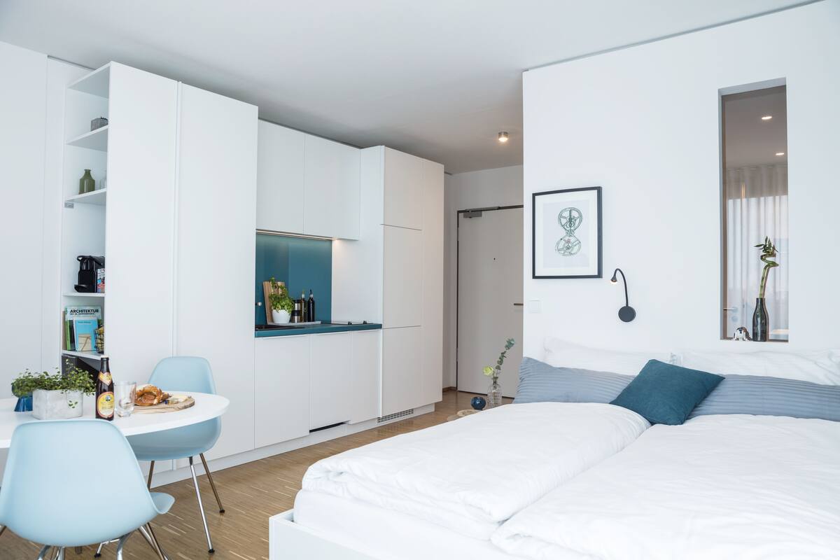A modern bedroom with a bed, a desk, and a chair. There is a small kitchenette area with a sink and a window.
