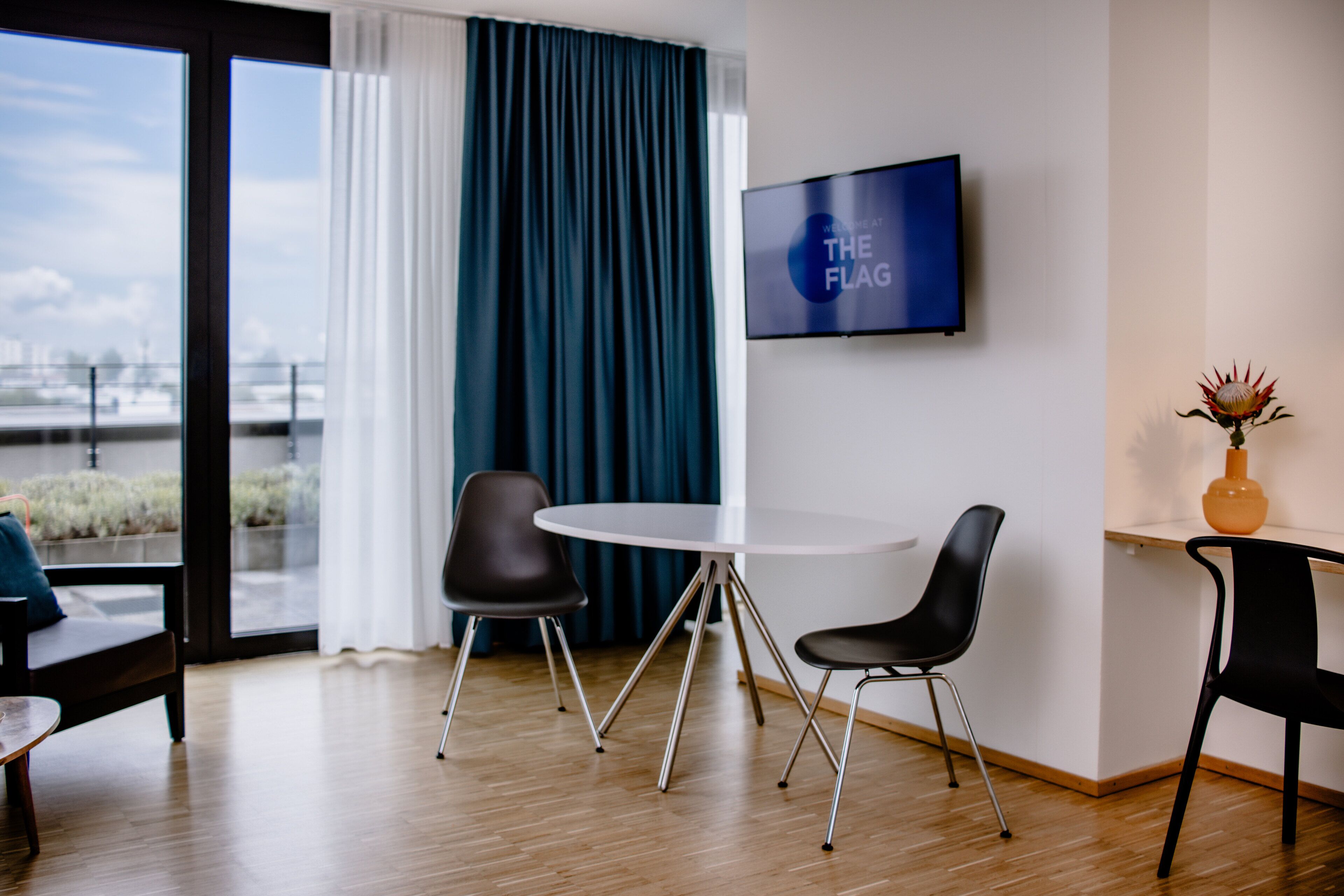 Una habitación de hotel moderna con un ventanal grande, una mesa redonda con dos sillas, un televisor de pantalla plana en la pared y un florero con una flor en una mesita de noche.