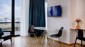 Una habitación de hotel moderna con un ventanal grande, una mesa redonda con dos sillas, un televisor de pantalla plana en la pared y un florero con una flor en una mesita de noche.