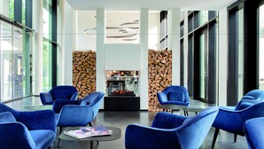 A modern lobby with blue chairs, a central table, and a fireplace. - THE FLAG München M. (Munich)