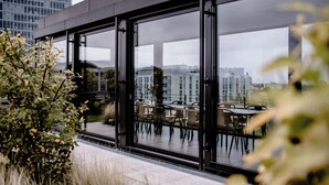 A modern outdoor seating area with glass doors, a view of the cityscape, and a mix of greenery. - THE FLAG München M. (Munich)