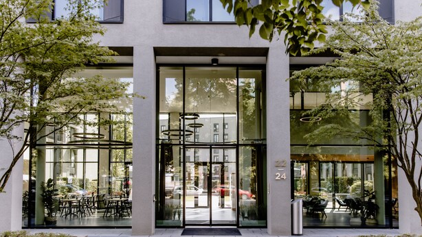 A modern building entrance with glass doors, outdoor seating, and a number '24' visible. - THE FLAG München M. (Munich)