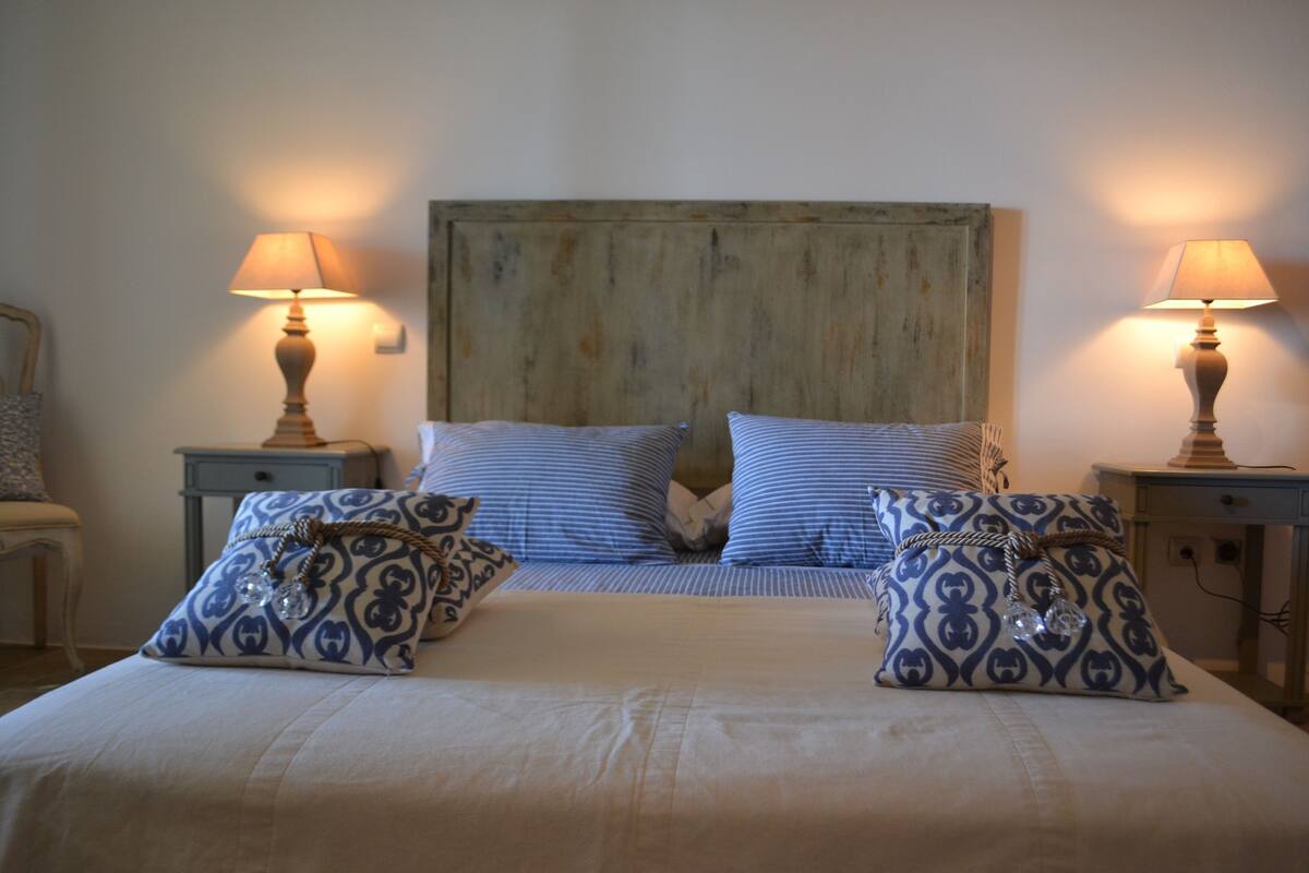Un lit bien fait, avec une literie à rayures et des coussins décoratifs, flanqué de deux tables de chevet avec des lampes, le tout adossé à une tête de lit en bois rustique.