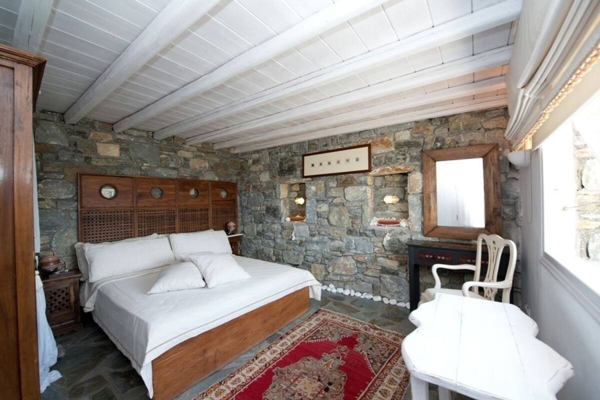Une chambre avec un mur en pierre, un lit en bois, un bureau blanc et une chaise.