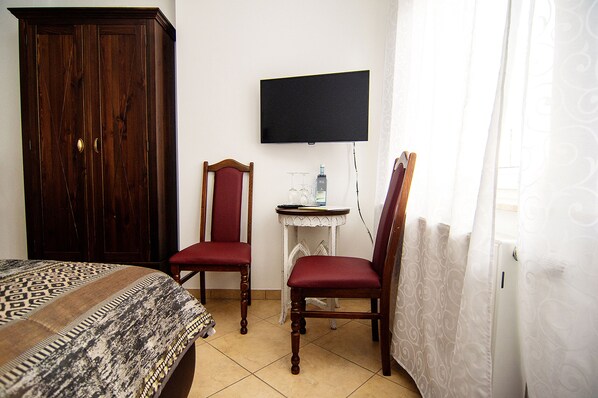 A hotel room with a bed, a wooden wardrobe, two red chairs, a small table with a bottle and glass, and a TV mounted on the wall.