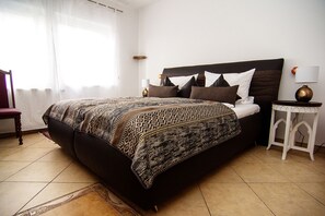 Una cama con una colcha estampada, una mesita de noche con una lámpara y una silla en una habitación con suelo de baldosas.