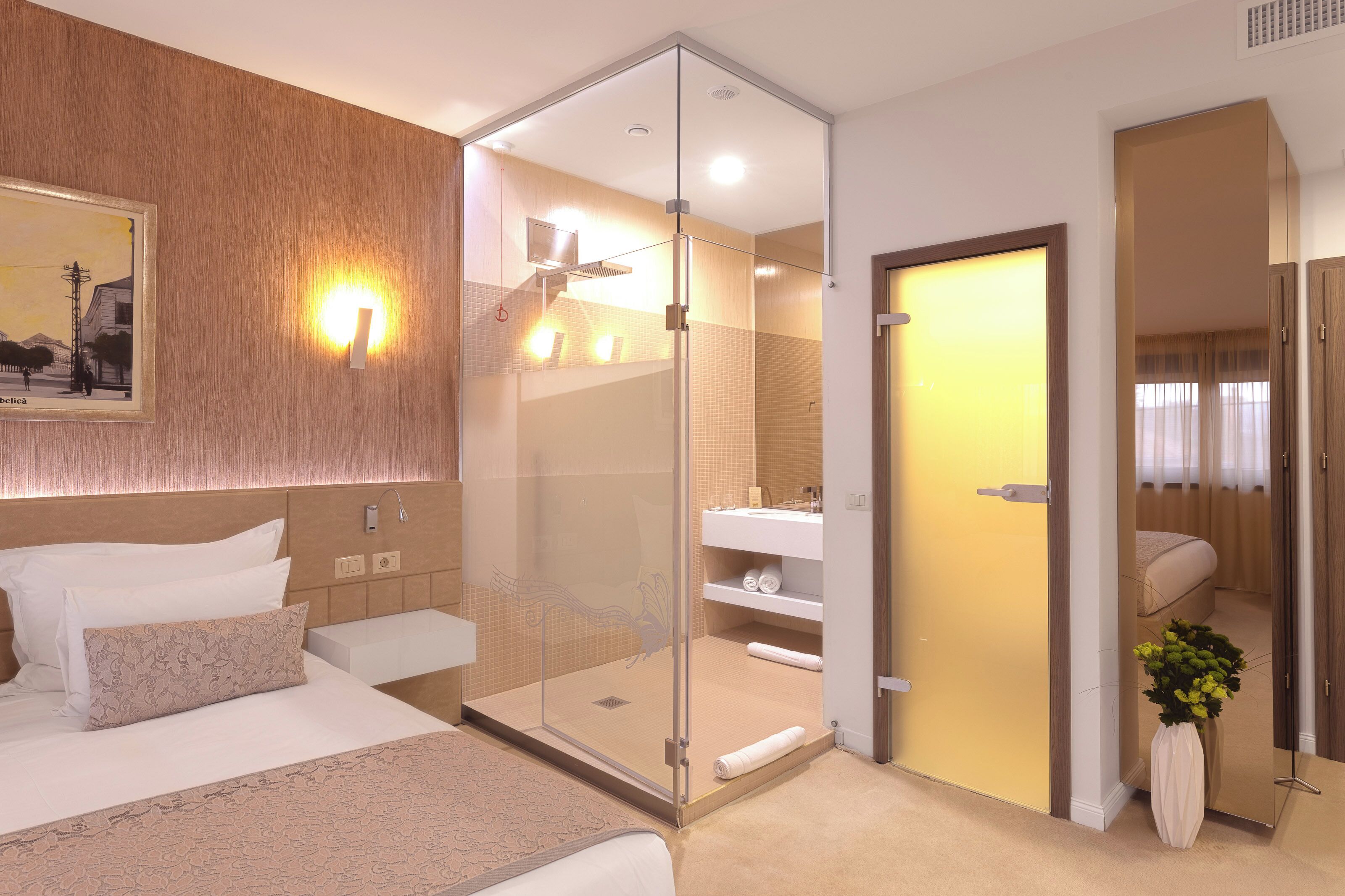 A modern hotel room with a bed, a glass-enclosed shower, a bathroom with a sink and mirror, and a painting on the wall.