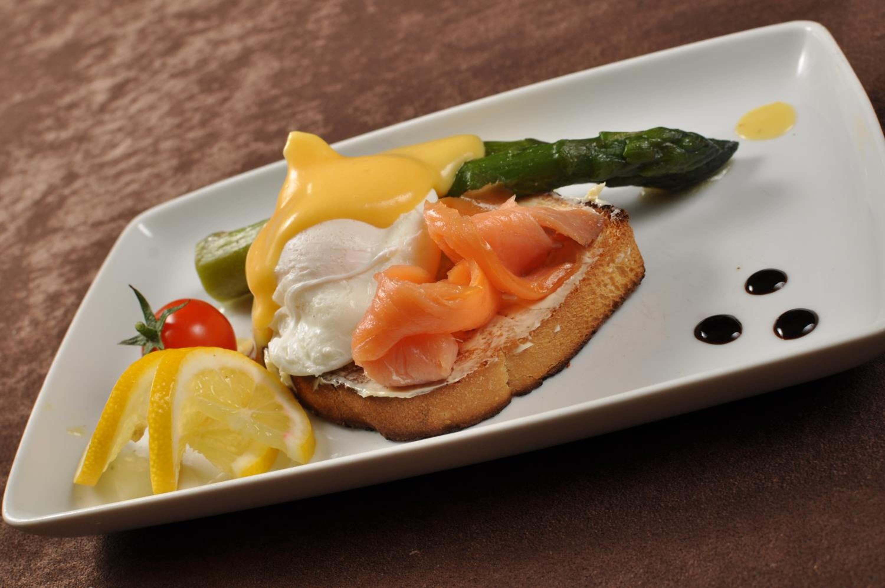 A plate with a sandwich, a slice of lemon, a cherry tomato, and a green vegetable, possibly asparagus, on a textured surface.