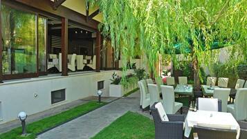 An outdoor patio area with tables and chairs, a pathway, and a building with large windows and a hanging willow tree.