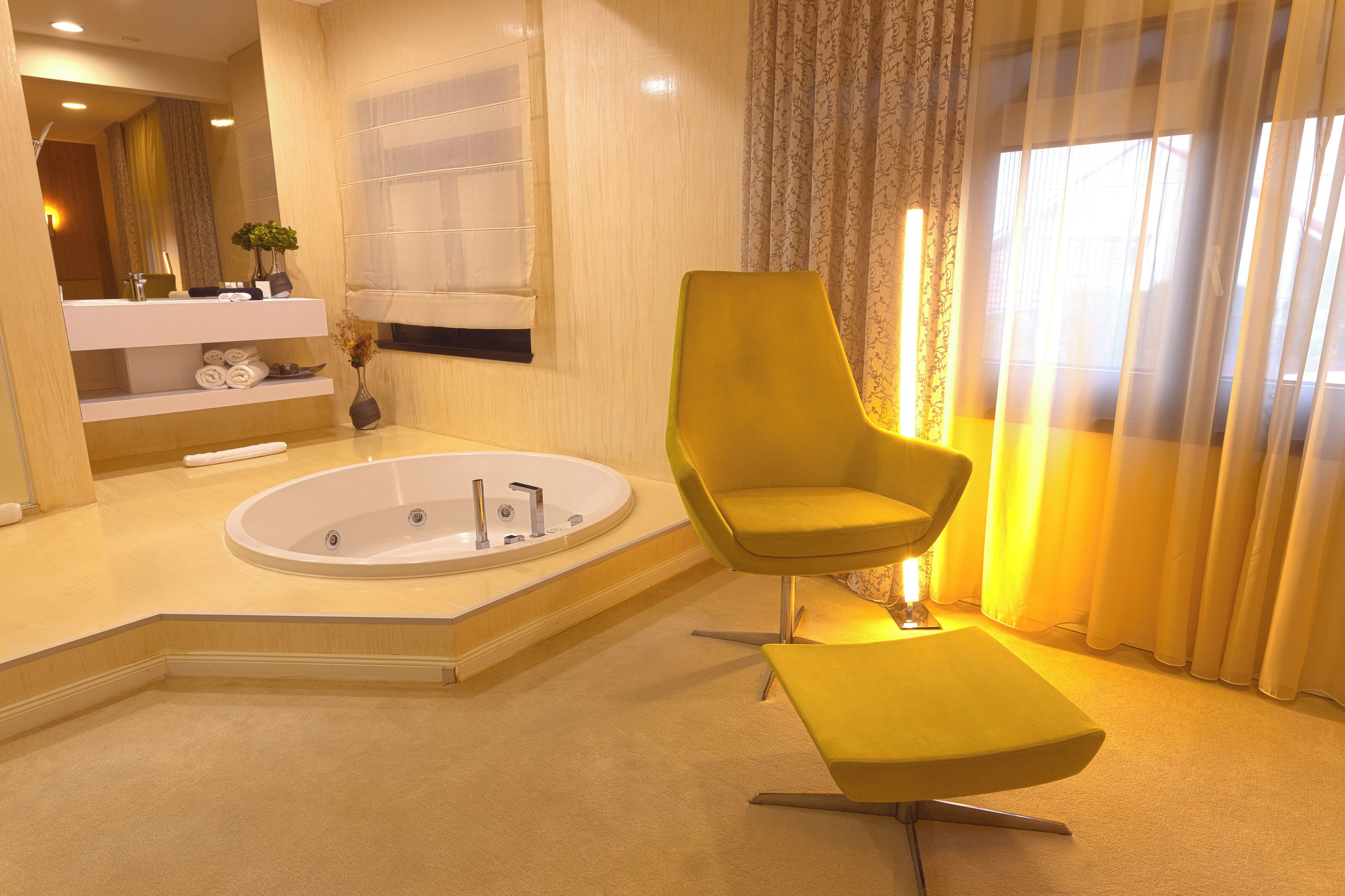 Un baño moderno con una bañera, una silla amarilla, un tocador con espejo y una ventana con cortinas.