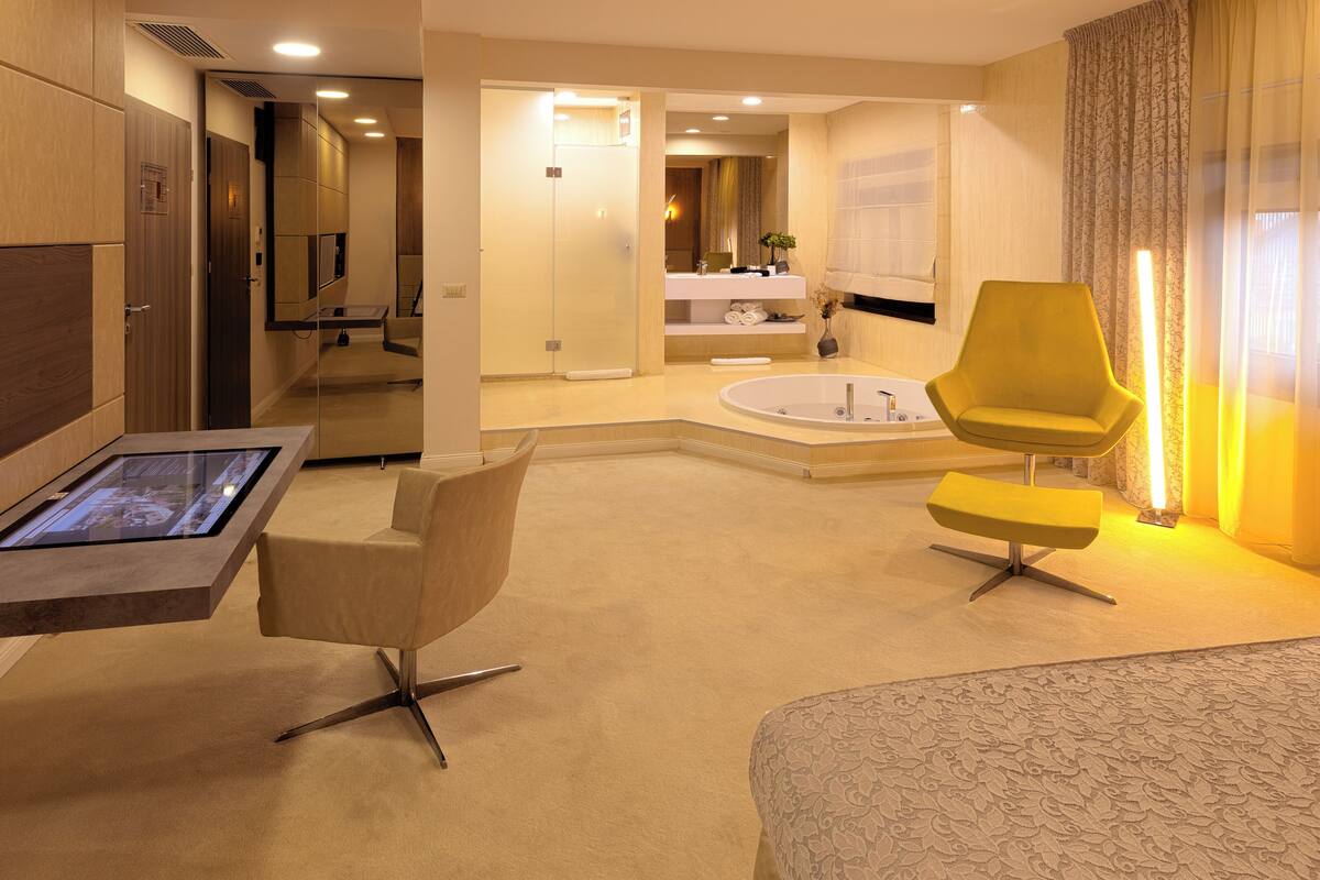 A modern hotel room with a large bed, a desk, a chair, a bathtub, and a mirror.