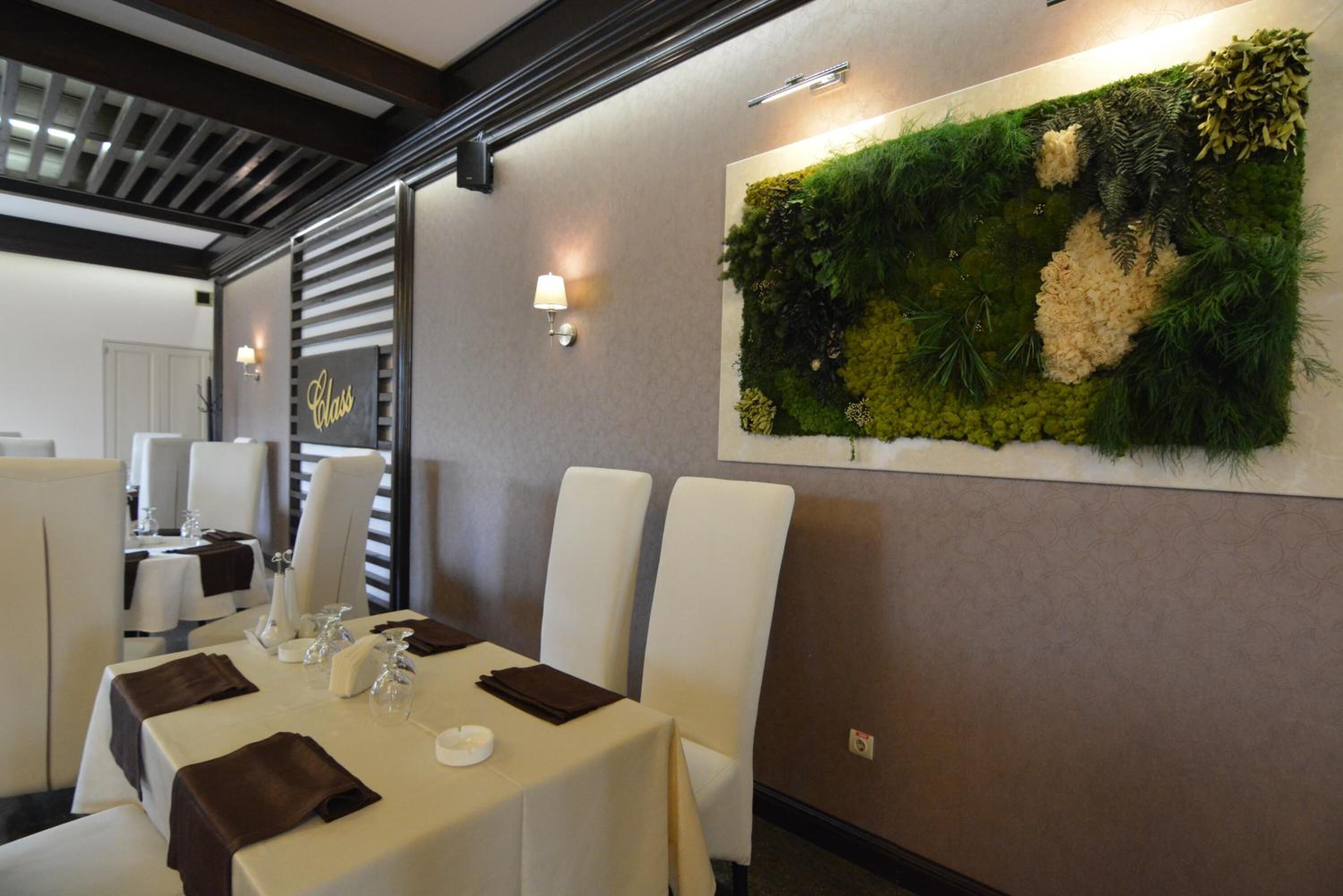 Un área de comedor con mesas y sillas blancas, una planta verde montada en la pared y un letrero que dice 'Glass'.