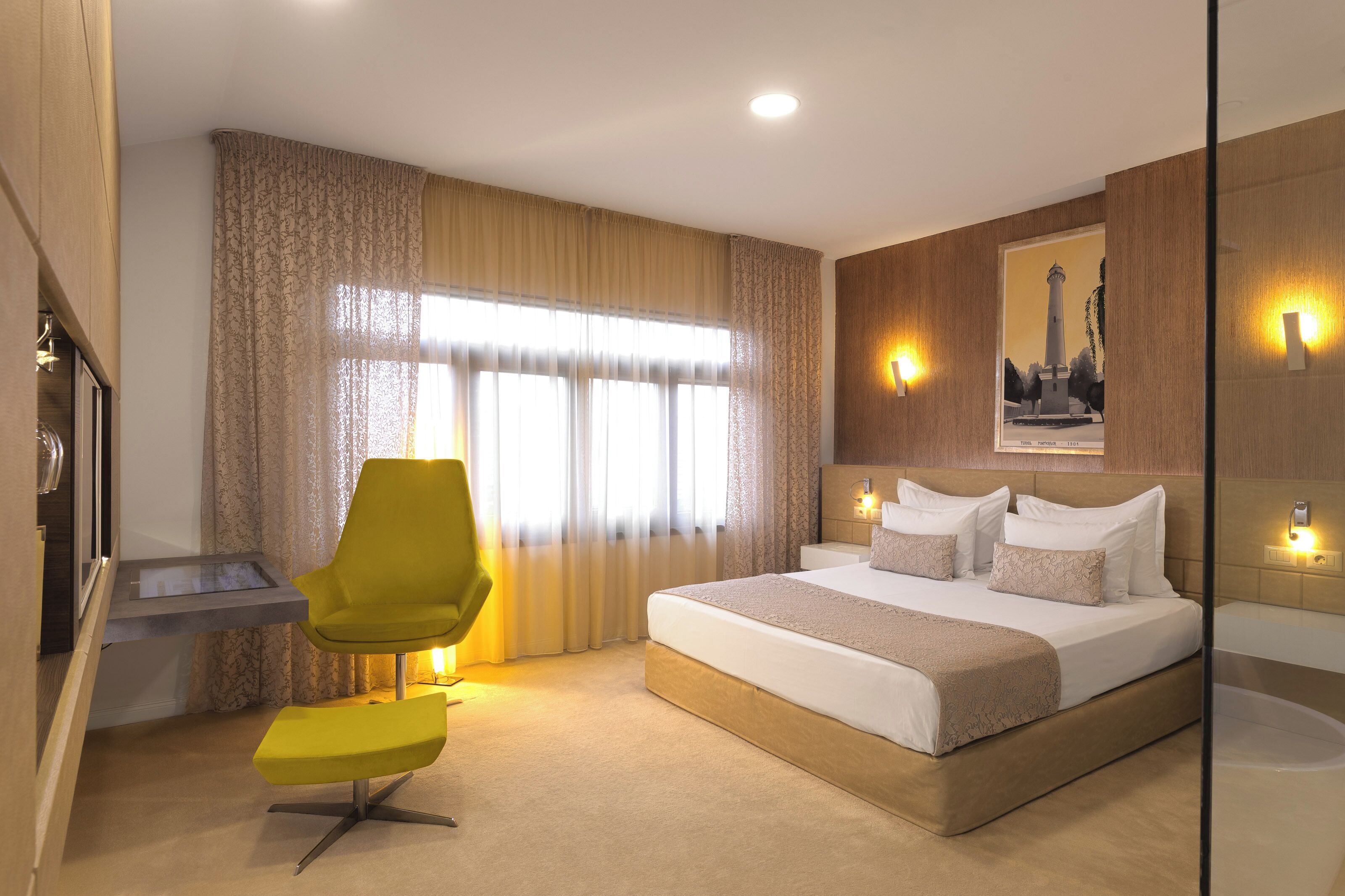A modern hotel room with a large bed, a desk, a chair, and a large window with curtains.