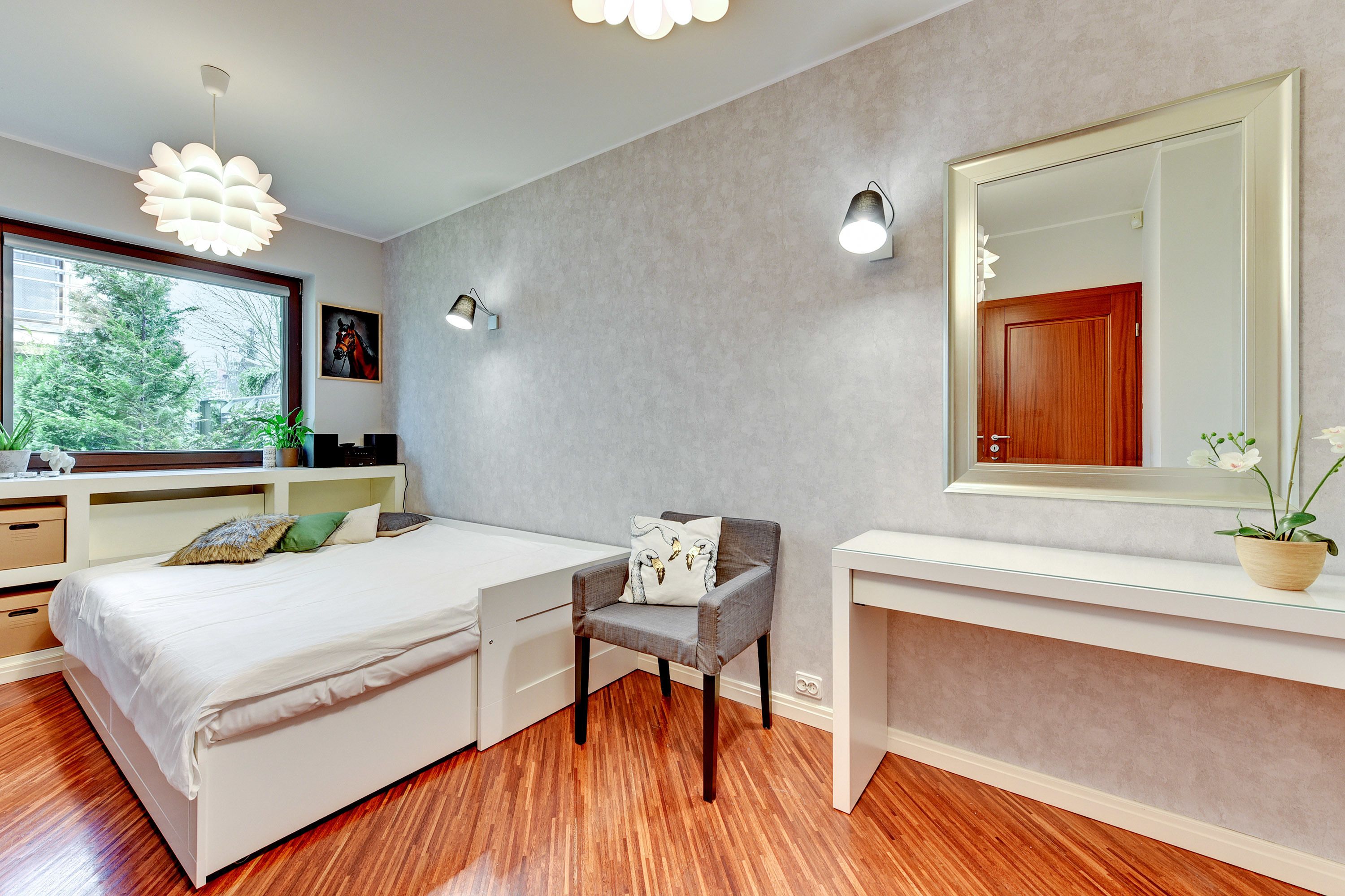A modern bedroom with a bed, a desk, a chair, a large mirror, and a window with a view of trees.