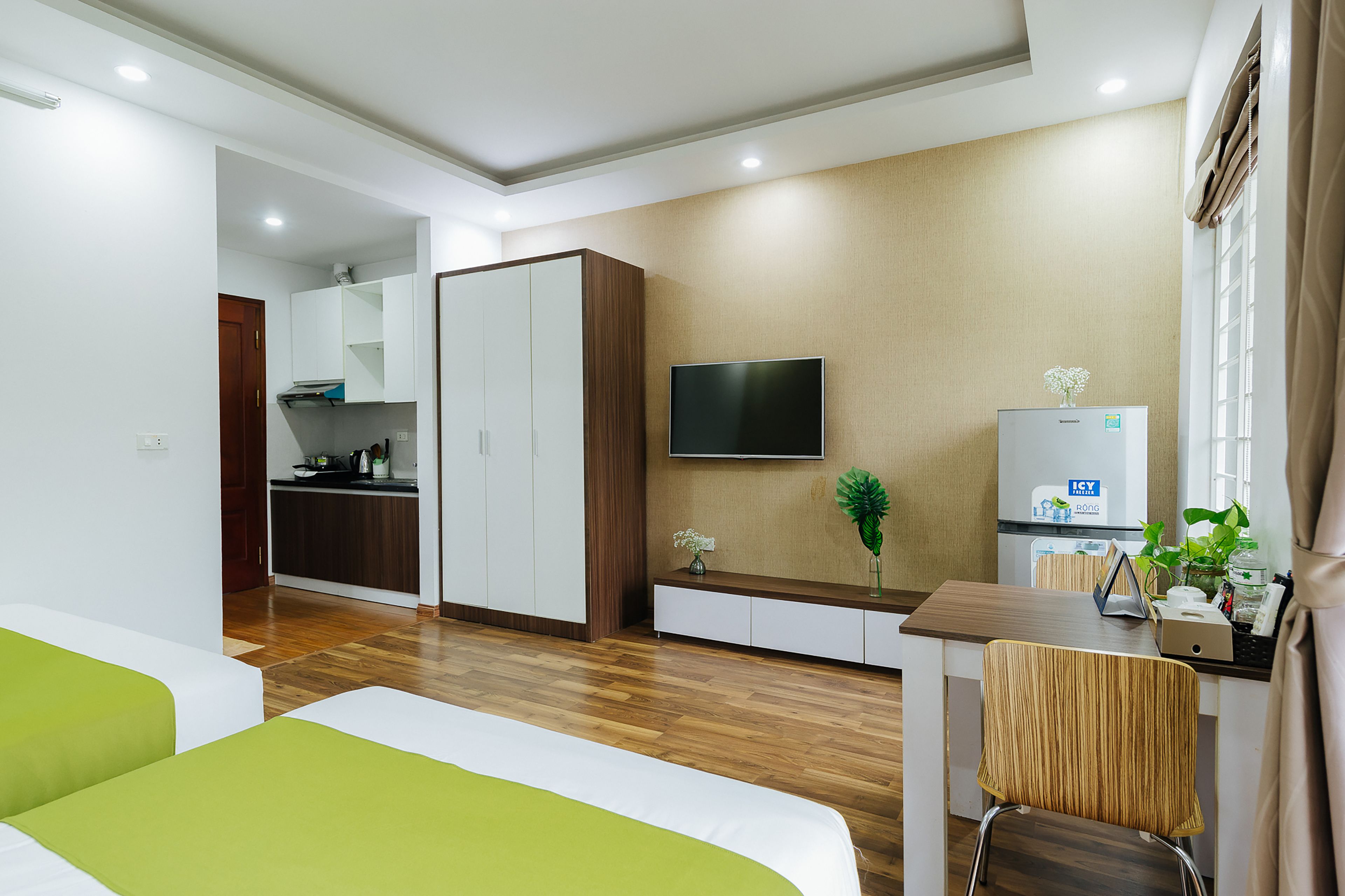 A modern hotel room with a bed, a desk with a chair, a TV, a kitchenette, and a bathroom.