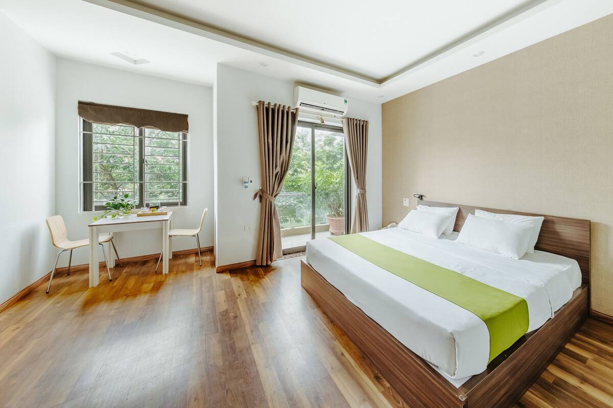 A hotel room with a wooden floor, a bed with white and green bedding, a desk with a chair, and a window with curtains.