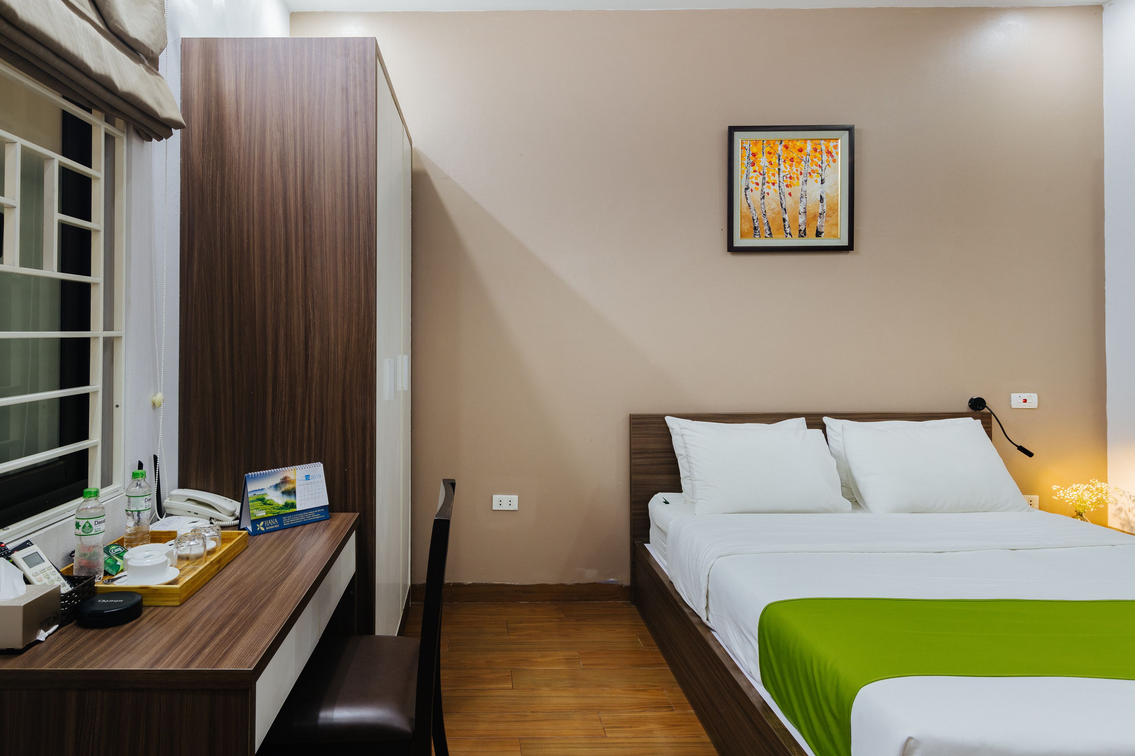 Habitación de hotel con cama, mesita de noche y escritorio con teléfono, bandeja con vasos y una botella, y un cuadro en la pared.