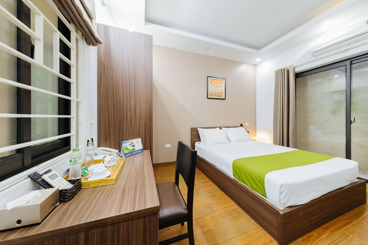 A hotel room with a wooden desk, a bed with a green bedspread, a window with blinds, and a painting on the wall.