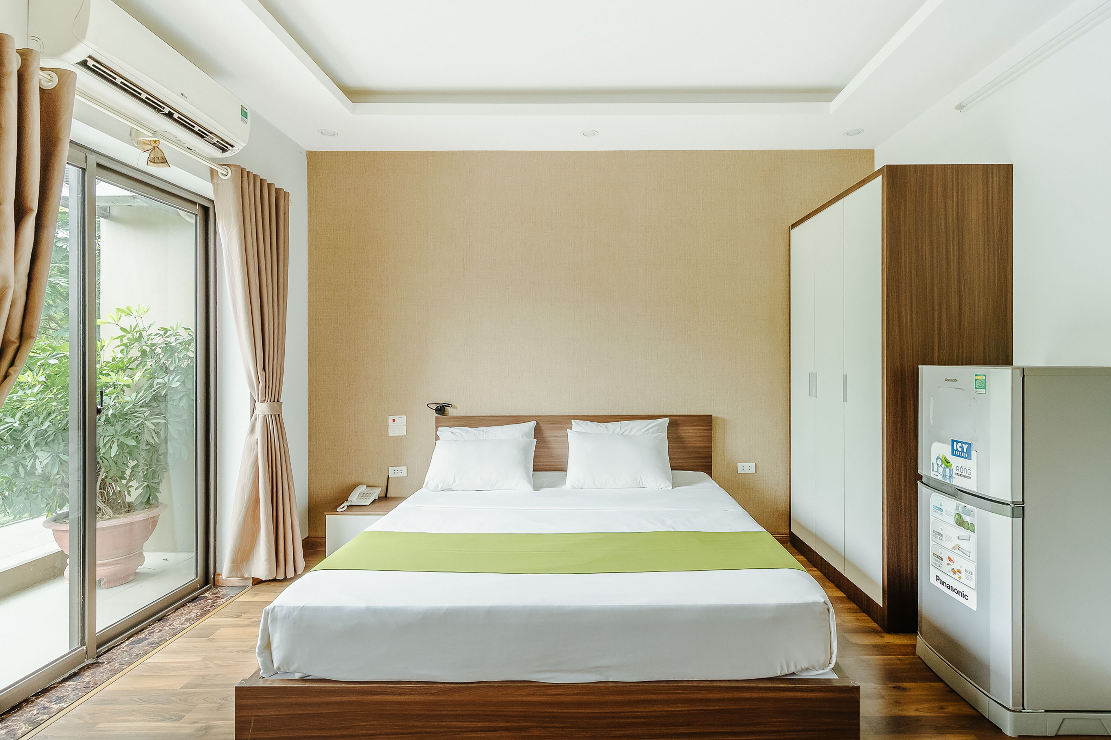 A hotel room with a bed, a nightstand, a refrigerator, and a sliding door to a balcony with a potted plant.