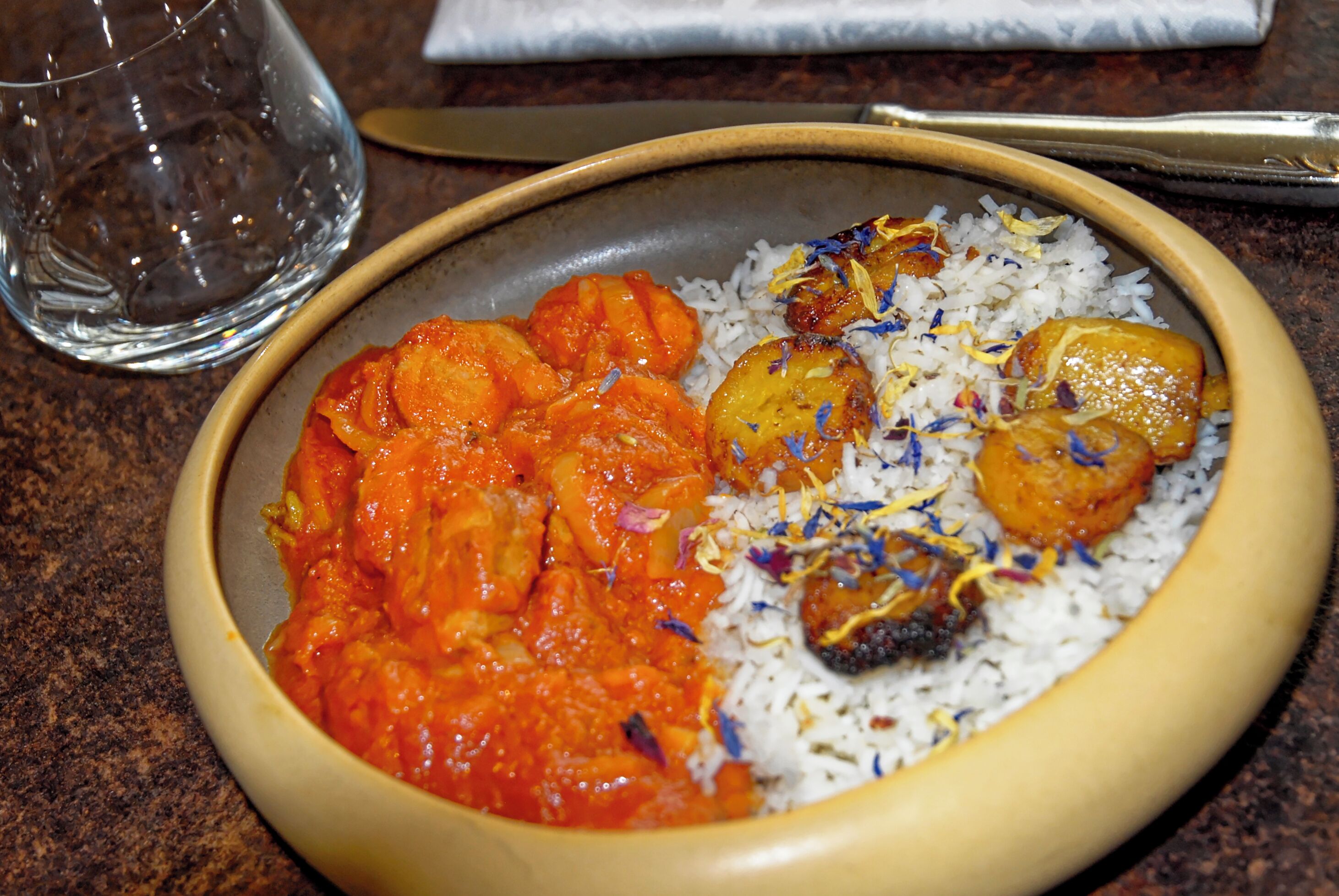 A bowl of food with rice and curry, a glass of water, and a spoon on a table.