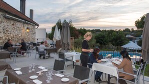Un área de comedor al aire libre con mesas preparadas para dos, un edificio de piedra al fondo y una piscina a la derecha.